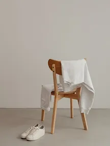 Wooden chair with a white shirt draped over its back and a pair of white sneakers on the floor beside it.