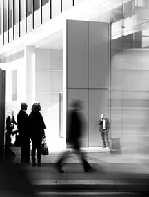 Black-and-white image of people walking and standing near a modern building with large glass windows.