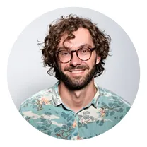 Smiling man with curly hair, beard, and glasses wearing a patterned shirt against a light gray background.
