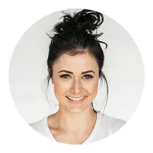 Smiling woman with dark hair in a messy bun wearing a white top against a light background.