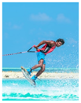 Man wearing a pink life jacket and blue shorts wakeboarding on turquoise water with blue sky background.
