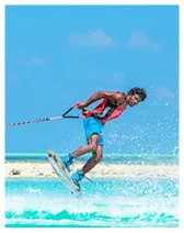 Man wearing a pink life jacket and blue shorts wakeboarding on turquoise water with blue sky background.