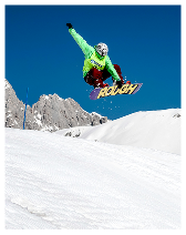 Snowboarder in green jacket and red pants airborne above snow-covered slope with mountains and clear blue sky.