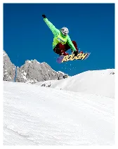 Snowboarder in green jacket and red pants airborne above snow-covered slope with mountains and clear blue sky.