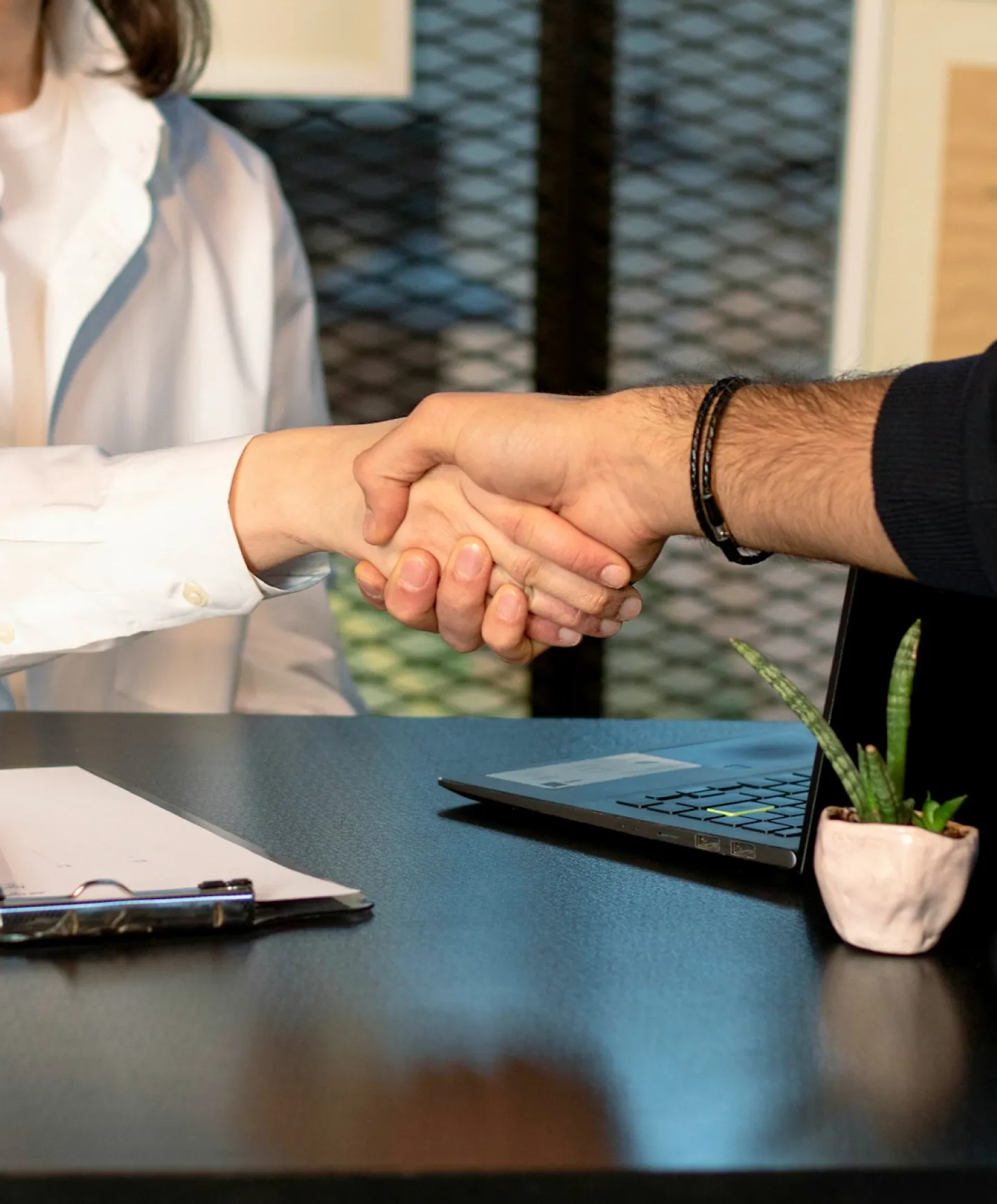 Two people shaking hands over a desk with a laptop, clipboard, and small potted plant.