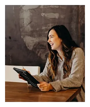 Young woman with long hair smiling and holding a notebook while sitting at a wooden table.