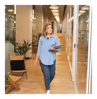 Woman in blue shirt and jeans walking in a modern office corridor holding a tablet and smiling.