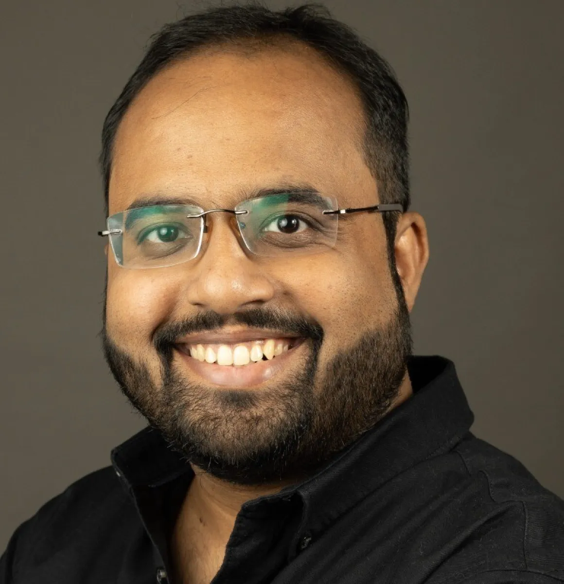 Smiling man with glasses and a beard wearing a black shirt against a neutral background.