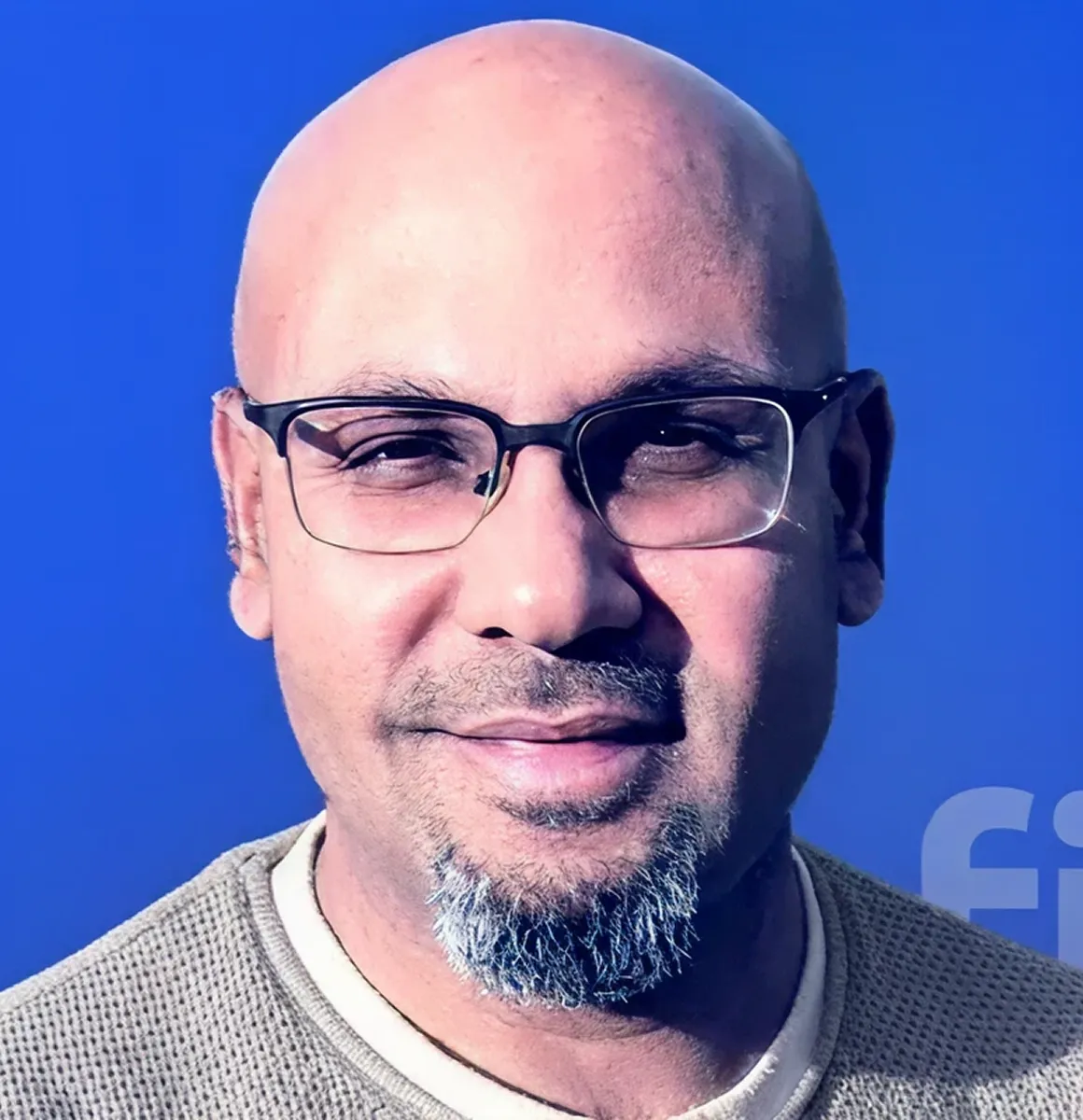 Close-up of a bald man with glasses, a salt-and-pepper goatee, and a beige sweater against a solid blue background.