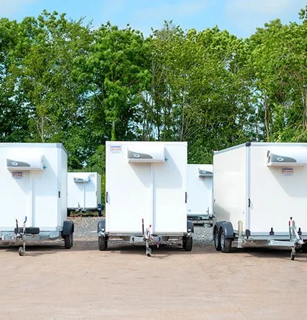 White refrigerated trailers parked outdoors in a row with green trees in the background under a partly cloudy sky.