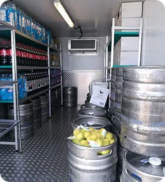 Interior of a commercial refrigerated storage with shelves holding bottles of water and soda, alongside metal kegs, some filled with lemons.