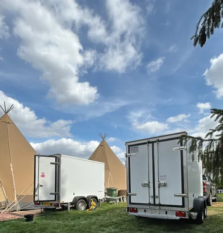 White trailers parked on grass in front of tan teepee tents under a blue sky with white clouds.