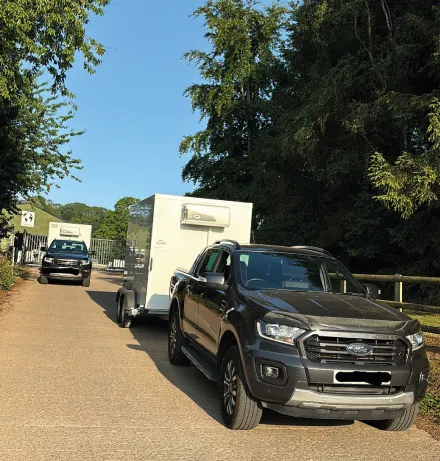 Gray Ford pickup truck towing a white enclosed trailer on a sunny road with another vehicle in the background and trees lining the roadside.