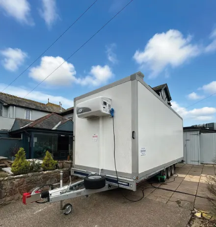 White refrigerated trailer parked outside on a paved area near buildings under a blue sky with clouds.