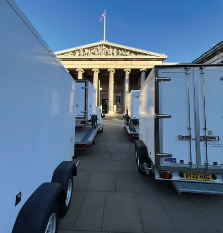 View between white trailers lined up on both sides leading to a classical building with columns and a British flag on top under clear blue sky.