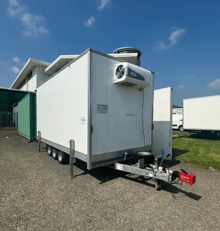 White refrigerated trailer with cooling unit parked on gravel near green and white buildings under a blue sky.