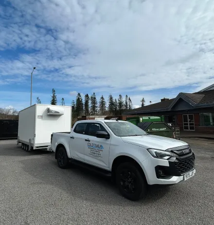 White pickup truck with ColdPalau branded door and attached refrigerated trailer parked on a paved area near buildings and trees.