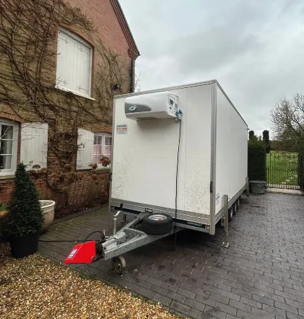 White refrigerated trailer parked on a driveway beside a brick house on a cloudy day.