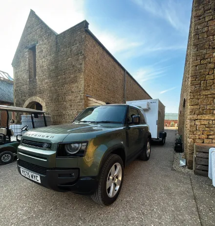 Green Land Rover Defender parked in a narrow stone courtyard next to historic brick buildings under a partly cloudy sky.
