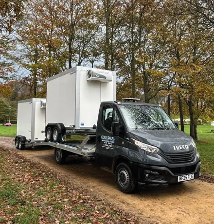 Black Iveco truck carrying a refrigerated trailer on a dirt path in a park with autumn trees.