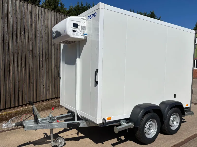White refrigerated trailer with dual axle and metal hitch parked beside a wooden fence.