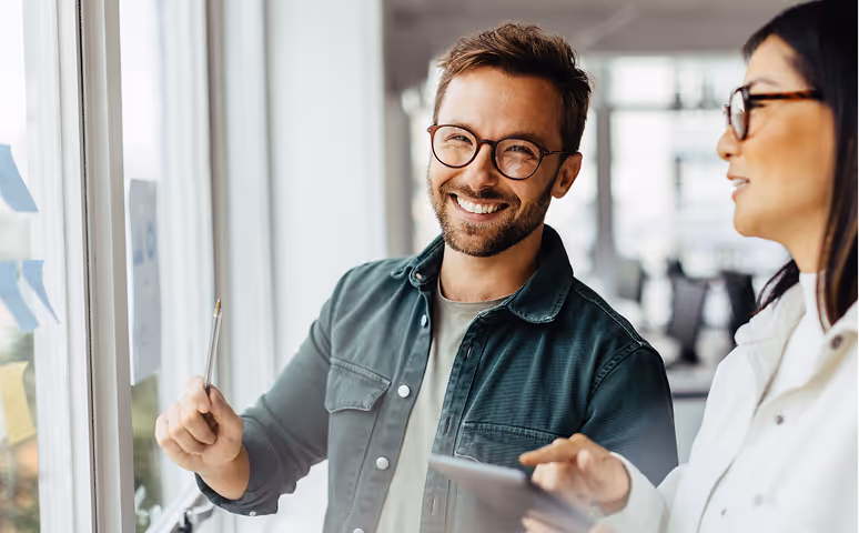 Twee collega's bespreken ideeën bij een raam met plakbriefjes, de man glimlacht terwijl hij een pen vasthoudt.