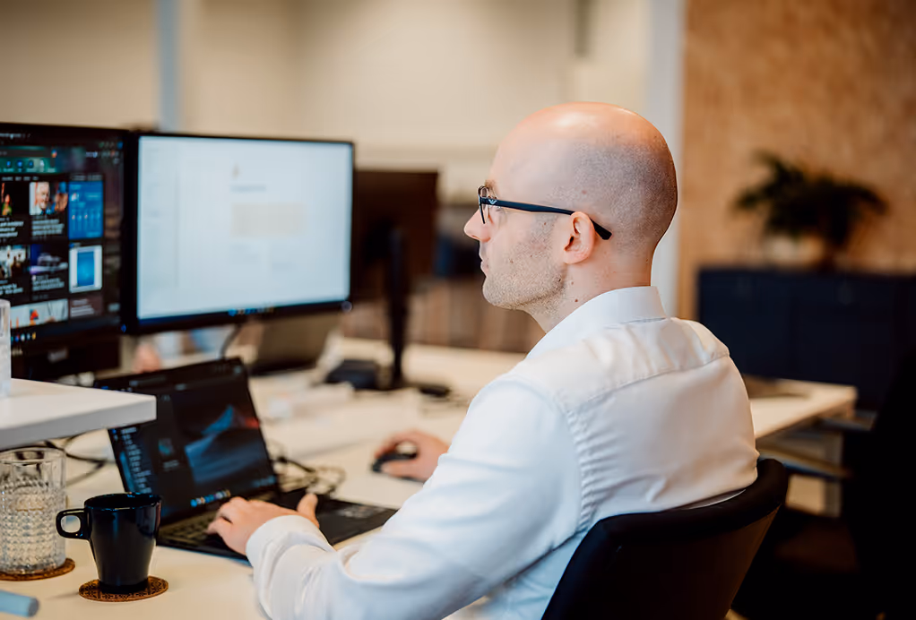 Man met bril en wit overhemd werkt op laptop en twee computerschermen in modern kantoor.