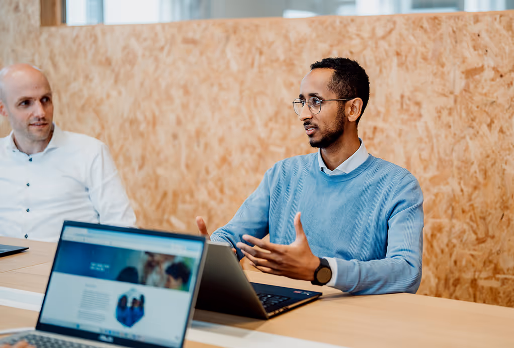 Twee mannen zitten aan een tafel en bespreken iets, een man in een blauwe trui spreekt terwijl de andere luistert.