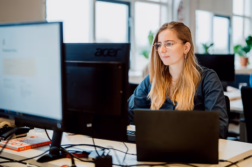 Vrouw met bril werkt op een laptop en desktop in een helder kantoor met planten aan het raam.