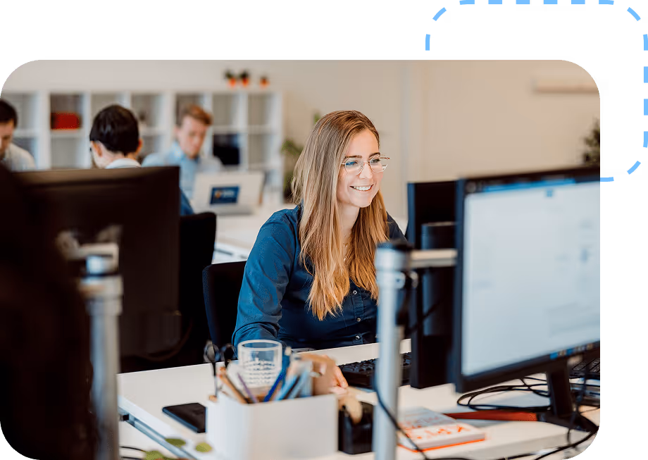 Vrouw met lichtbruin haar en bril glimlacht terwijl ze aan een bureau werkt met een computer in een kantooromgeving.