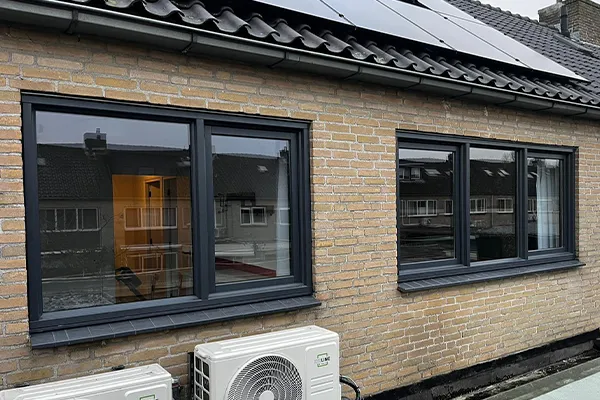 Twee grote zwarte kunststof kozijnen in een gevel van een woning, met weerspiegeling van overbuurhuizen en airco-units onder het raam.