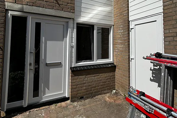 Witte kunststof voordeur met naastgelegen kozijn en opbergdeur, in een hoek van een bakstenen gevel met wit rabat