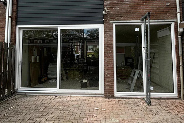 Witte kunststof schuifpui en groot raam in een bakstenen gevel, met zicht op een verbouwing binnen