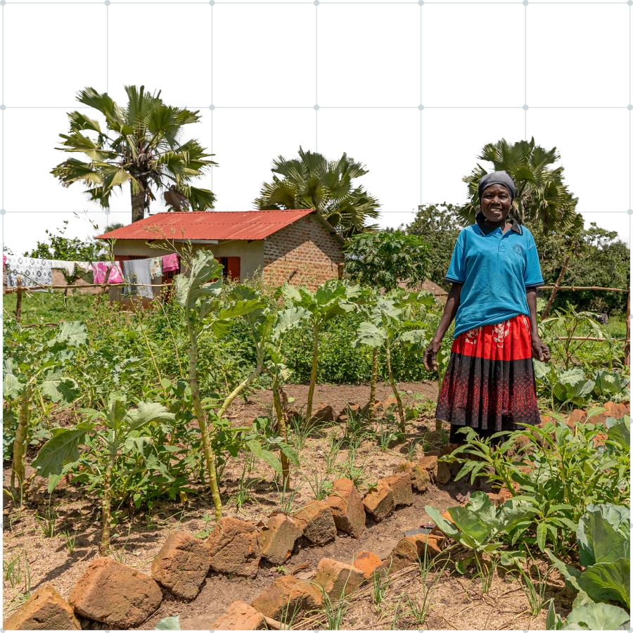 Woman wearing a blue shirt and red skirt standing in a garden with various plants and a brick house with a red roof in the background.