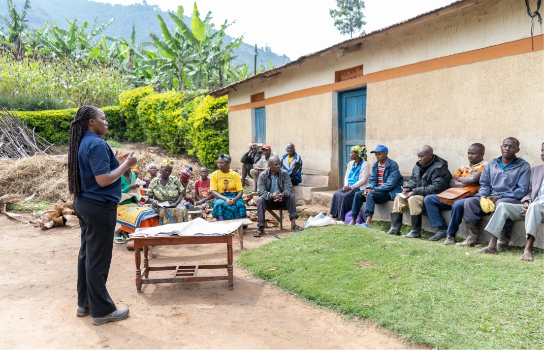 Woman standing and speaking to a group of people seated outdoors near a building in a rural setting.