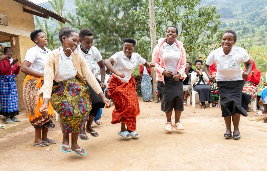 Group of women joyfully dancing outdoors on a dirt ground with green trees and hills in the background.