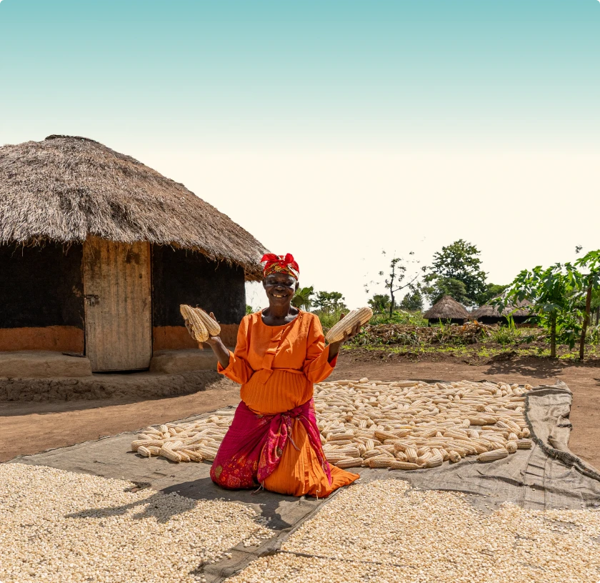 Smiling woman in orange and red traditional clothing kneeling outdoors with dried corn cobs and kernels spread on mats near a thatched hut.