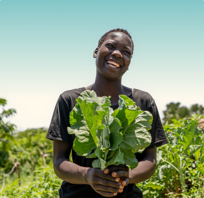 Smiling person holding fresh leafy greens in a sunny garden or farm setting.