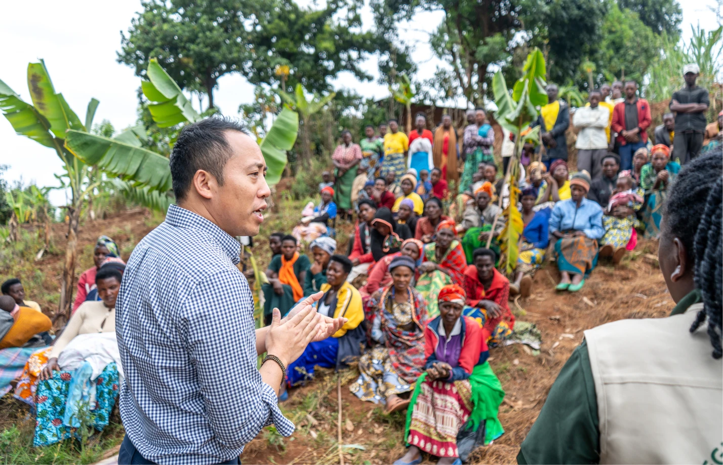 Man in checkered shirt speaking to a seated group of people outdoors in a rural area with banana plants.