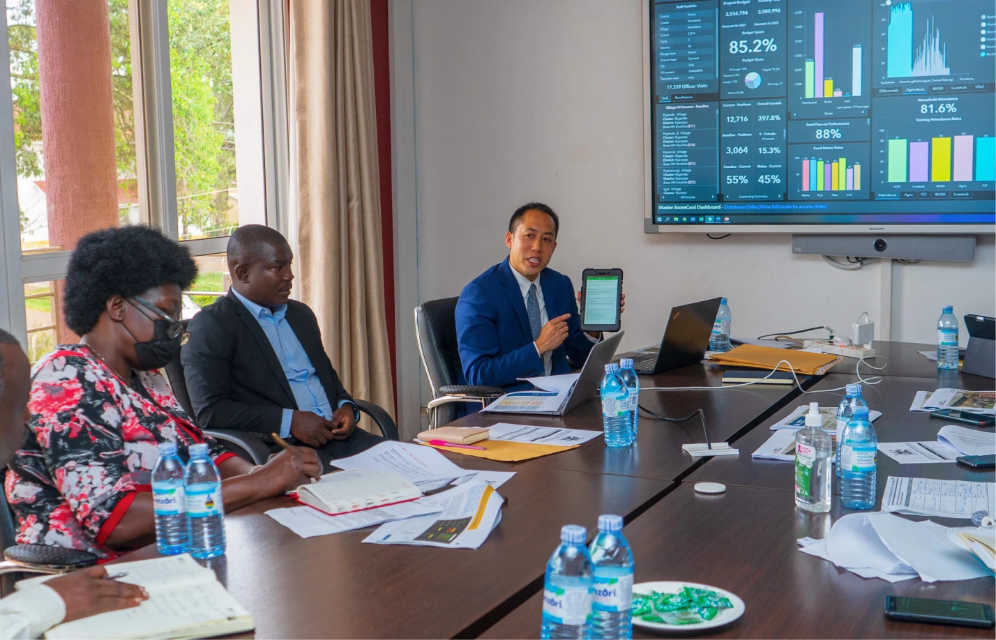A man in a blue suit presenting data on a tablet during a meeting with colleagues seated at a conference table with laptops, documents, water bottles, and a large screen showing charts.