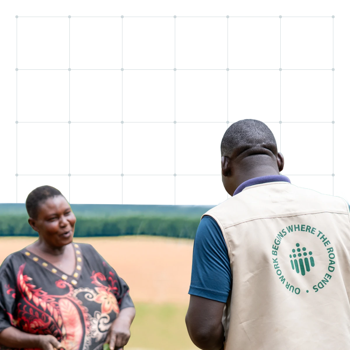 Two women drawing water from a hand pump in a fenced rural area with green hills and scattered trees in the background.