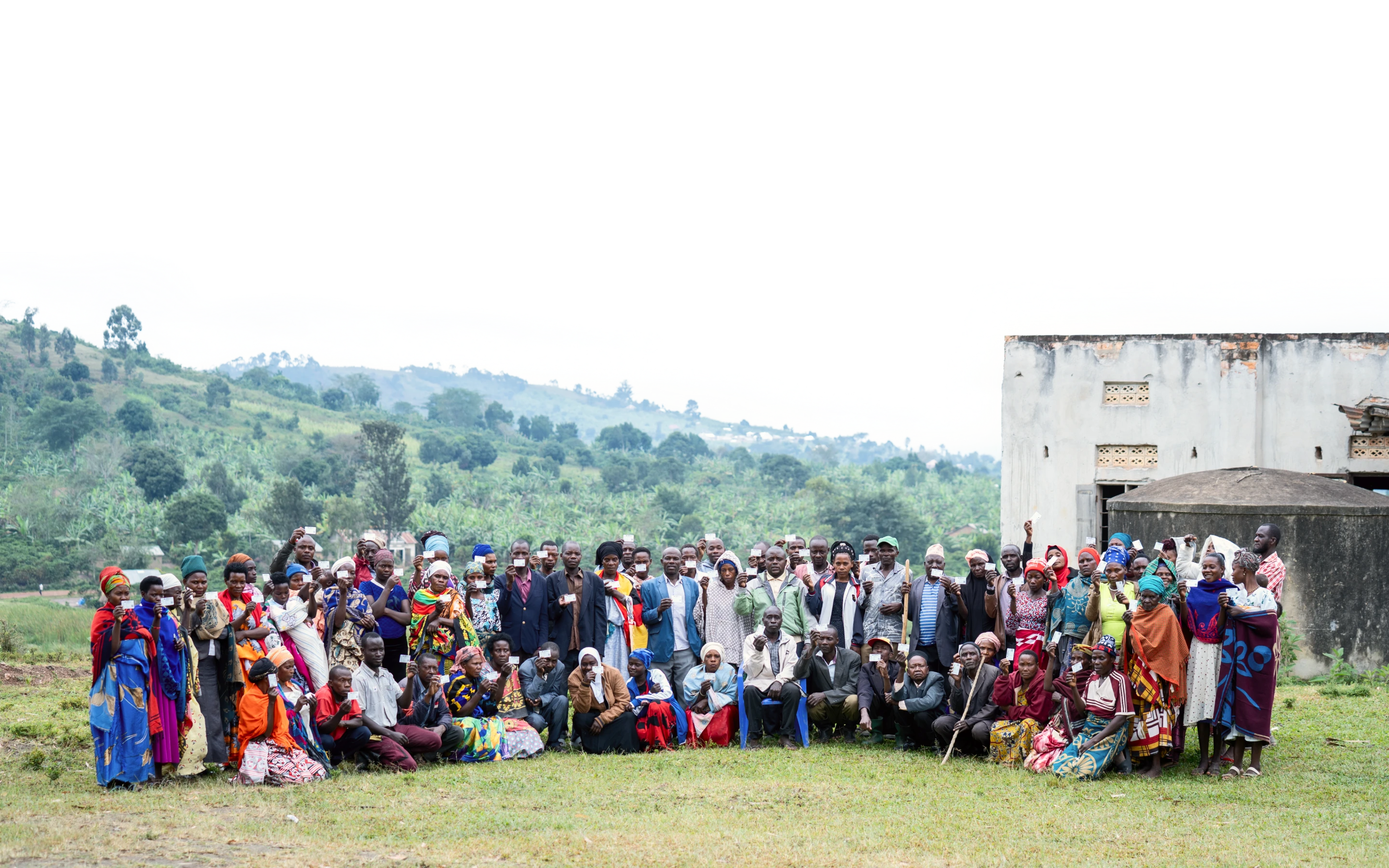 Large group of people posing outdoors in front of greenery and a building, many holding up identity cards.