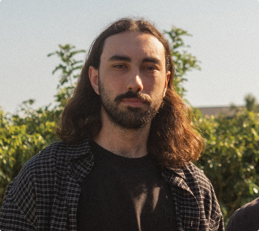 Man with long brown hair, beard, and mustache wearing a black shirt and a black and white checkered shirt standing outdoors with greenery and a clear sky in the background.