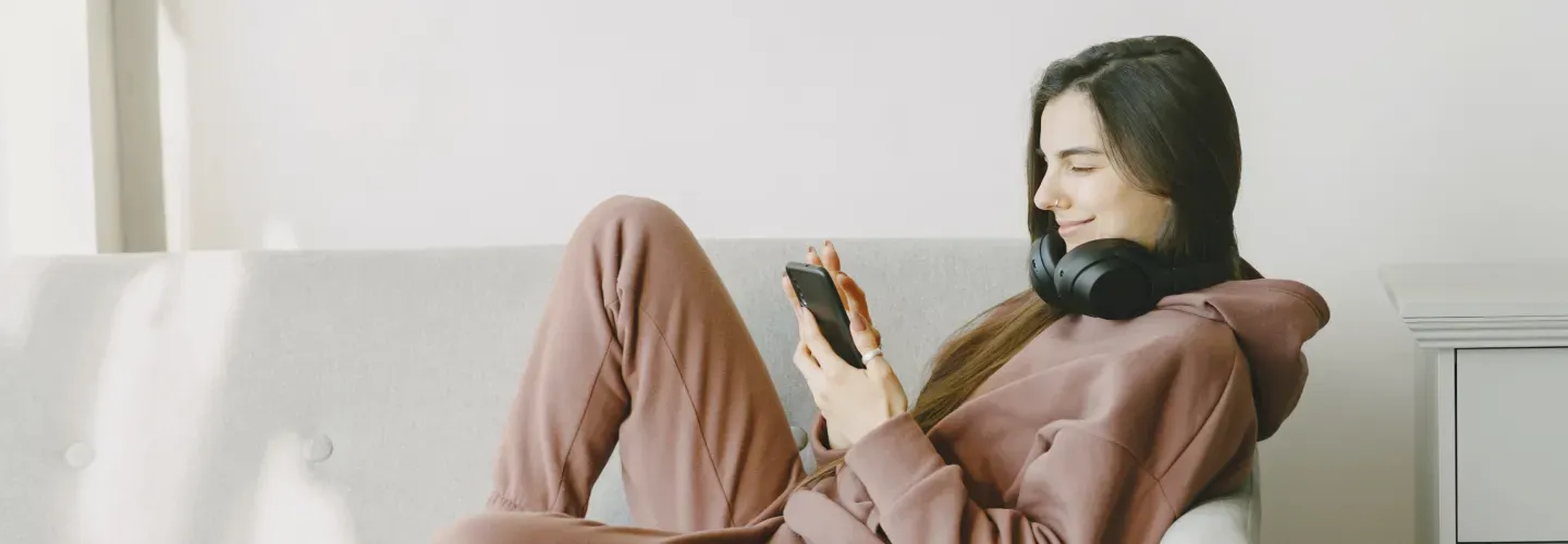 Mujer sonriente sentada en un sofá gris con auriculares negros alrededor del cuello mirando su teléfono móvil.