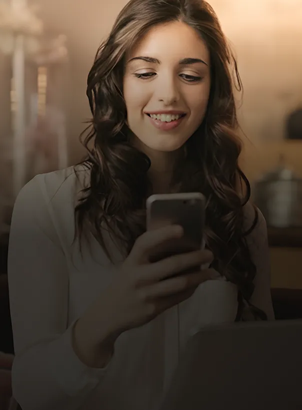 Mujer sonriente mirando su teléfono móvil, sentada frente a una computadora portátil en un ambiente interior.