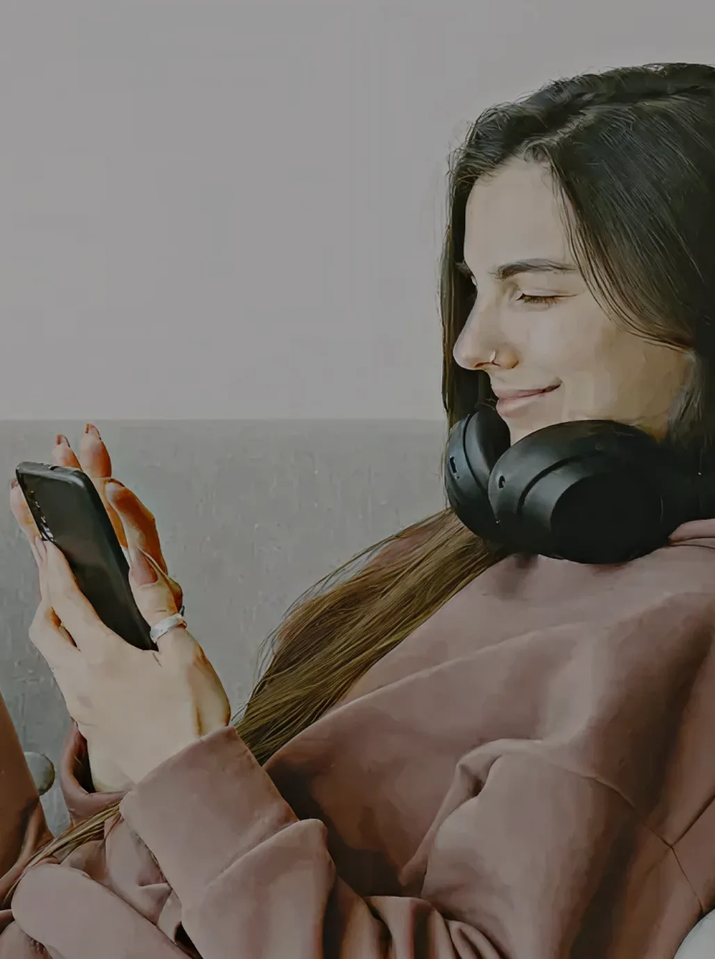Mujer sonriente sentada en un sofá gris con auriculares negros alrededor del cuello mirando su teléfono móvil.