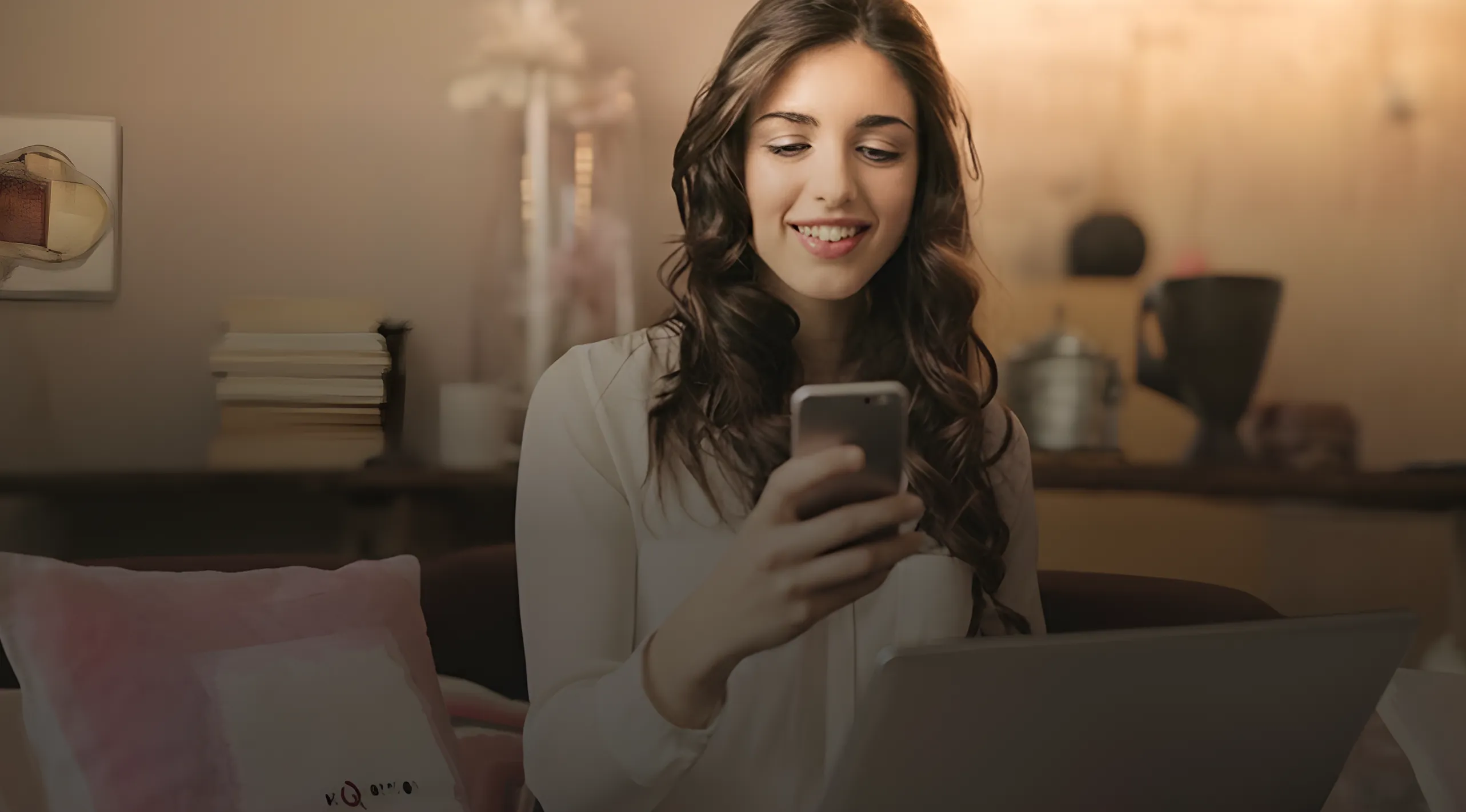 Mujer joven sonriente usando un teléfono móvil y con una computadora portátil en un entorno doméstico.
