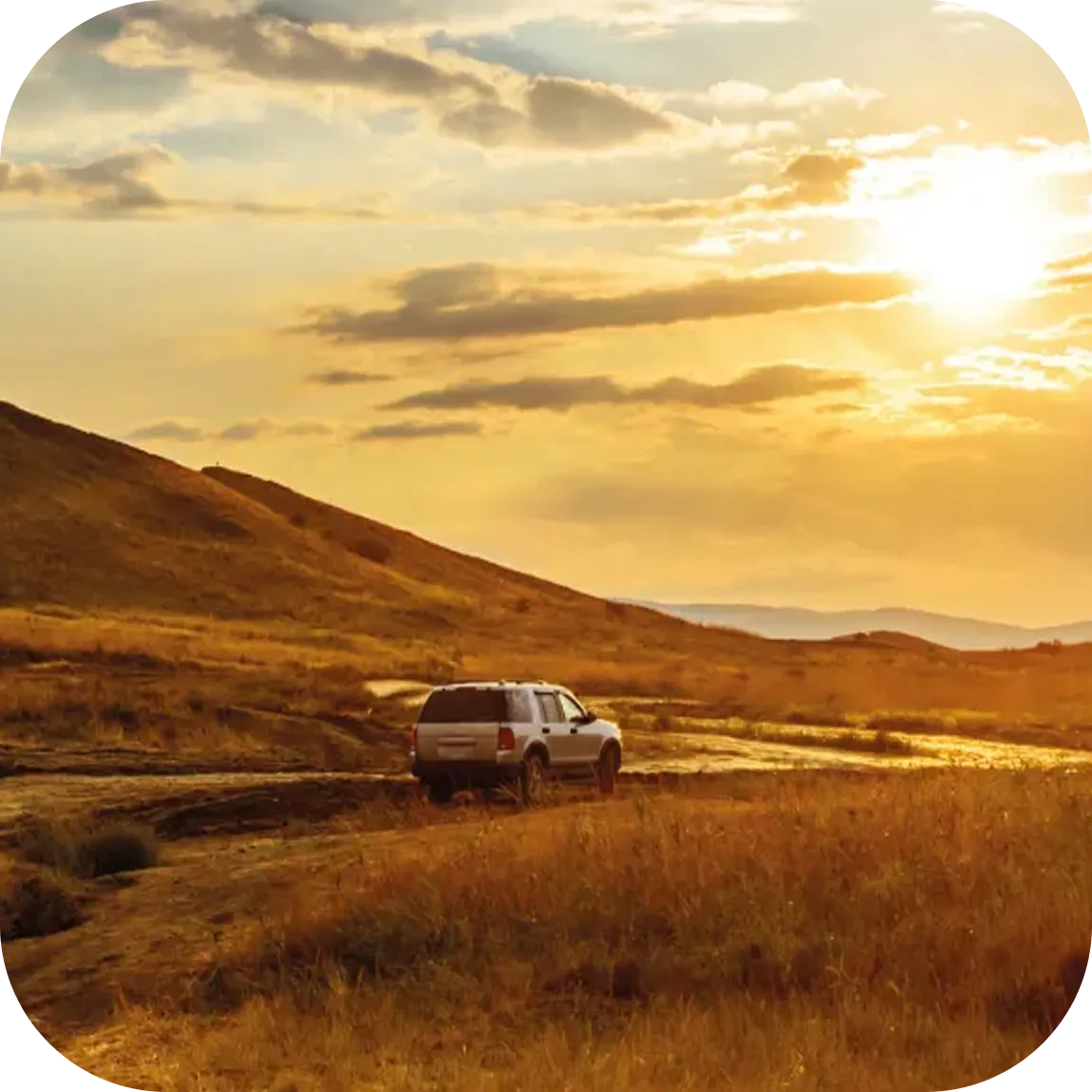 Un SUV blanco conduciendo por un camino de tierra en un paisaje rural de colinas bajo un cielo al atardecer.