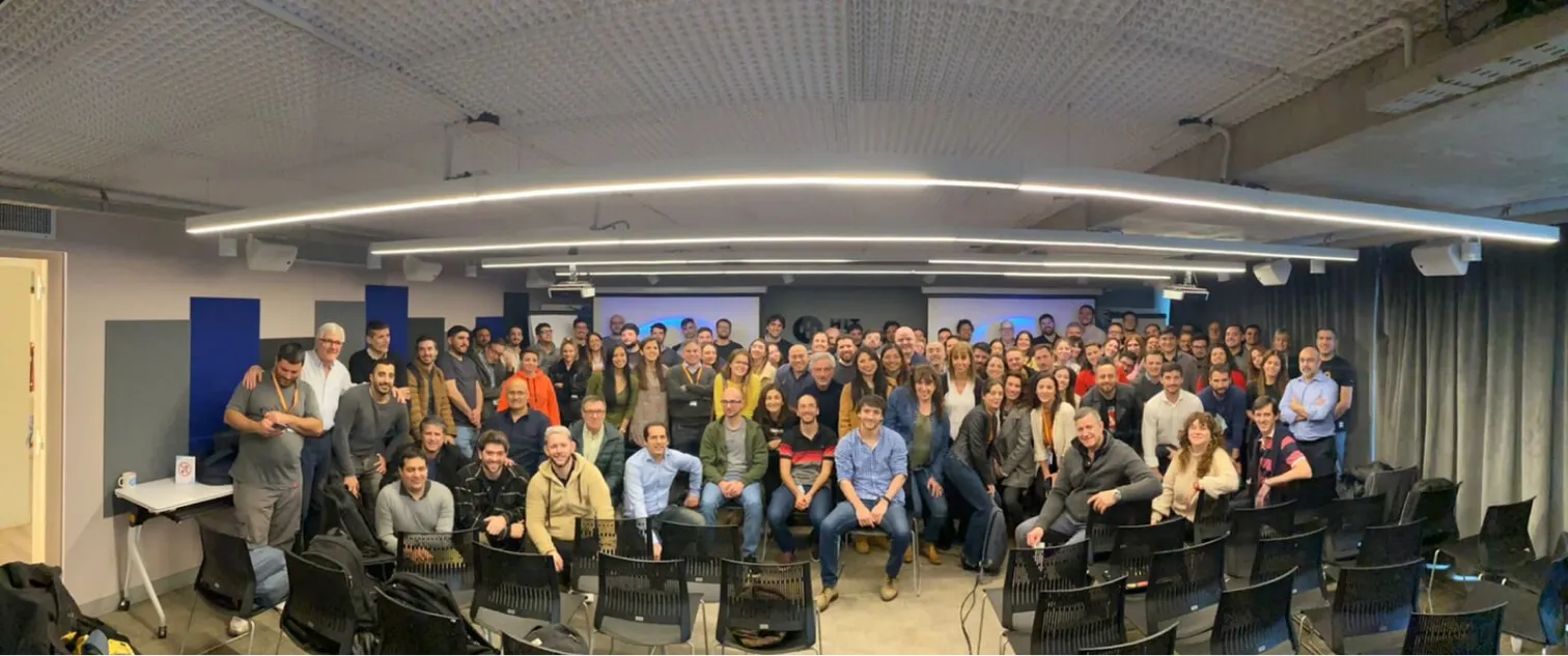 Gran grupo de personas en una sala de reuniones moderna con luces curvas en el techo y varias sillas vacías al frente.