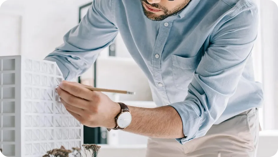 Hombre adulto ajustando un modelo arquitectónico blanco con un lápiz en una oficina luminosa.